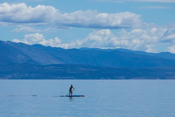 Una persona hace pádel surf en un mar en calma, con montañas y nubes al fondo