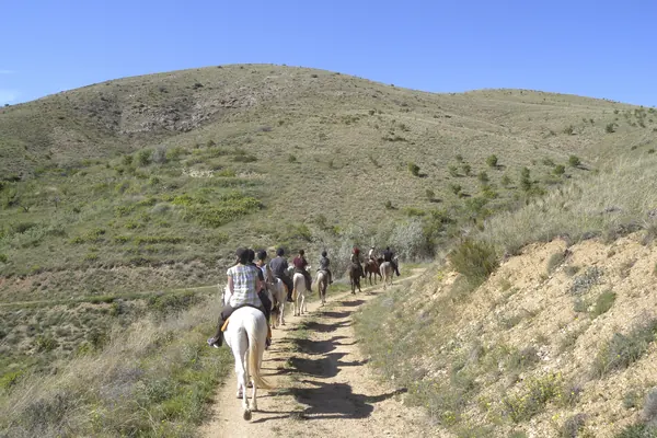 Un grupo a caballo por un camino de tierra entre colinas, visto de espaldas (foto genérica, no es Gavà)