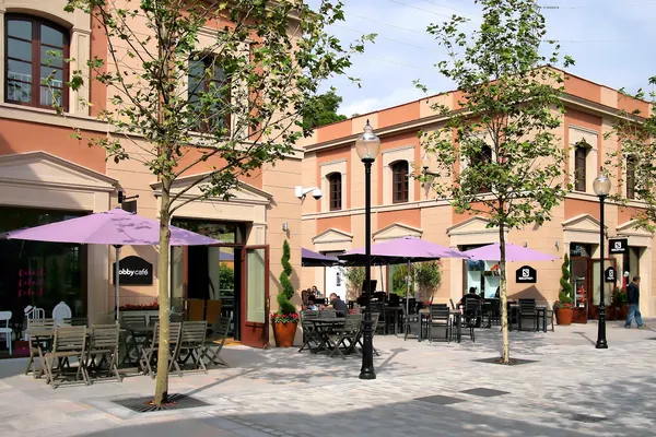 Una calle de La Roca Village con terrazas, sombrillas lilas y árboles, entre edificios de colores