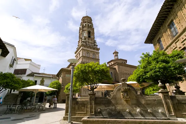 Una plaza del Poble Espanyol con su fuente de piedra y la torre mudéjar, bajo un cielo azul