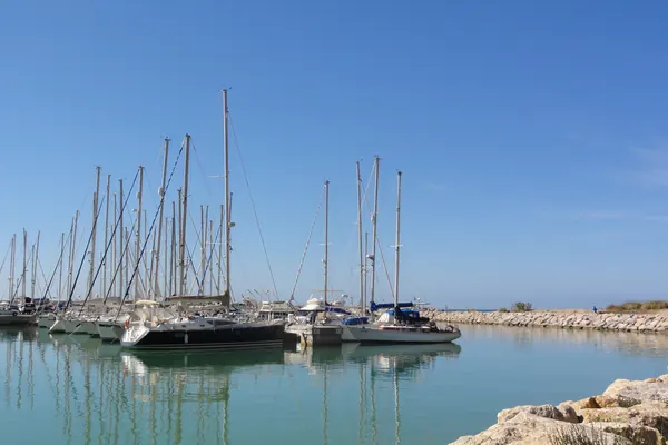 Veleros amarrados en Port Ginesta reflejados en el agua tranquila del puerto, bajo un cielo azul